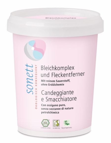 Das Bild zeigt einen weißen Behälter mit rosa Etikett der Marke Sonett. Auf dem Etikett steht 'Bleichkomplex und Fleckenentferner'. Der Behälter hat ein weißes, abnehmbares Oberteil. Auf dem Etikett sind auch Logos für ökologische Zertifizierungen sichtbar.