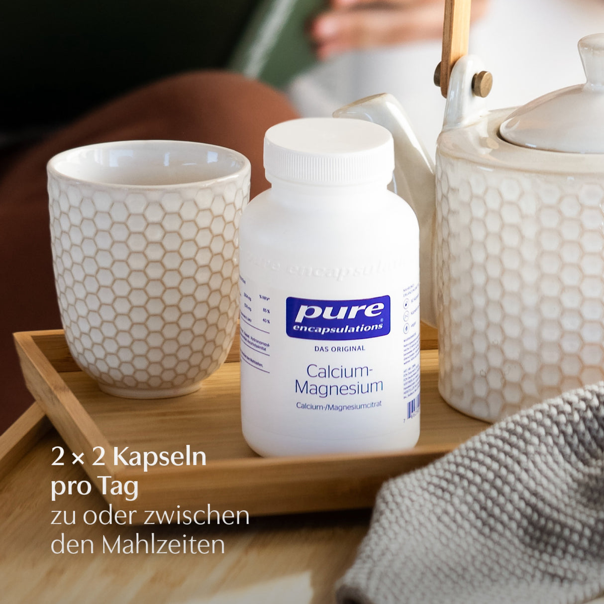 Das Bild zeigt eine weiße Flasche mit der Aufschrift 'pure encapsulations Calcium-Magnesium' auf einem Holztablett. Daneben stehen eine weiße Tasse und eine Teekanne mit Wabenmuster. Im Hintergrund ist eine Person unscharf zu erkennen. Auf dem Bild steht der Text: '2 x 2 Kapseln pro Tag zu oder zwischen den Mahlzeiten'.