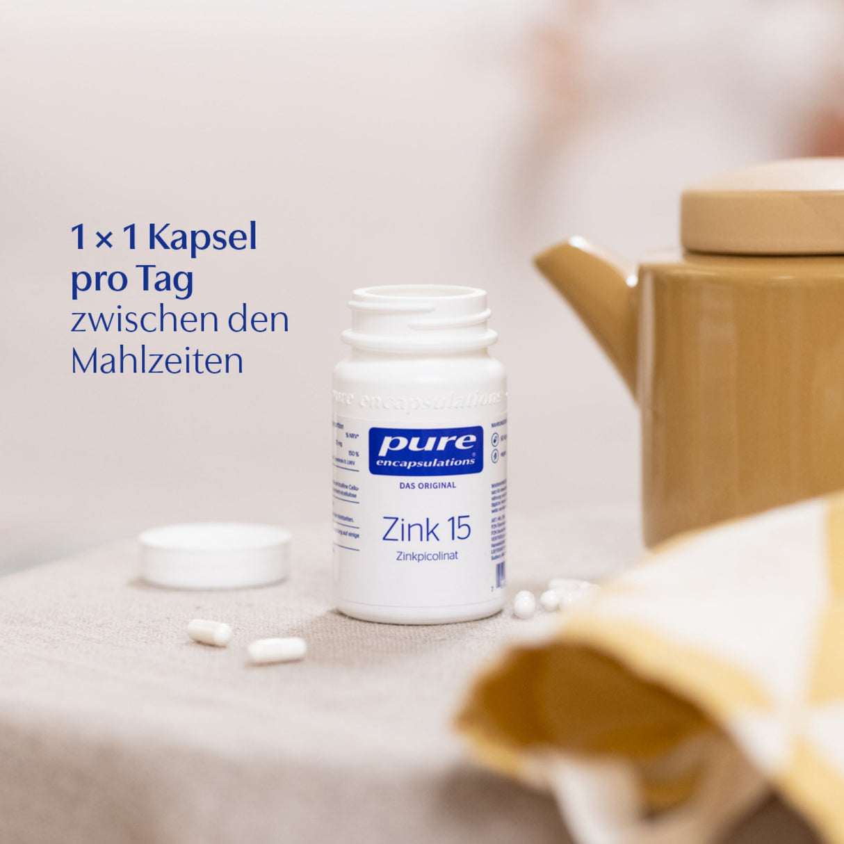 Das Bild zeigt eine weiße Flasche mit der Aufschrift 'pure encapsulations Zink 15 Zinkpicolinat' auf einem Tisch. Daneben liegt ein weißer Deckel und einige Kapseln. Im Hintergrund ist eine beige Teekanne zu sehen. Auf der linken Seite steht der Text '1 × 1 Kapsel pro Tag zwischen den Mahlzeiten'.