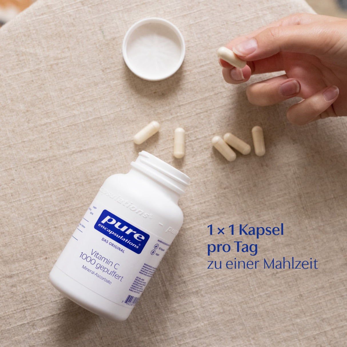 Auf einem beigen Tisch liegt eine geöffnete weiße Flasche mit der Aufschrift 'pure encapsulations Vitamin C 1000 gepuffert'. Daneben liegen mehrere Kapseln. Eine Hand greift nach einer Kapsel. Ein Text auf dem Bild sagt: '1 x 1 Kapsel pro Tag zu einer Mahlzeit'.