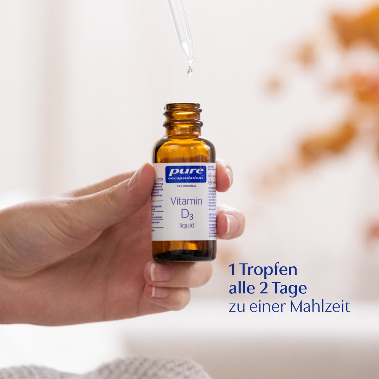 Eine Hand hält eine kleine braune Glasflasche mit der Aufschrift 'Vitamin D3 liquid' von Pure Encapsulations. Über der Flasche befindet sich eine Pipette, aus der ein Tropfen fällt. Im Hintergrund ist unscharf eine helle Umgebung zu sehen. Auf dem Bild steht der Text: '1 Tropfen alle 2 Tage zu einer Mahlzeit'.