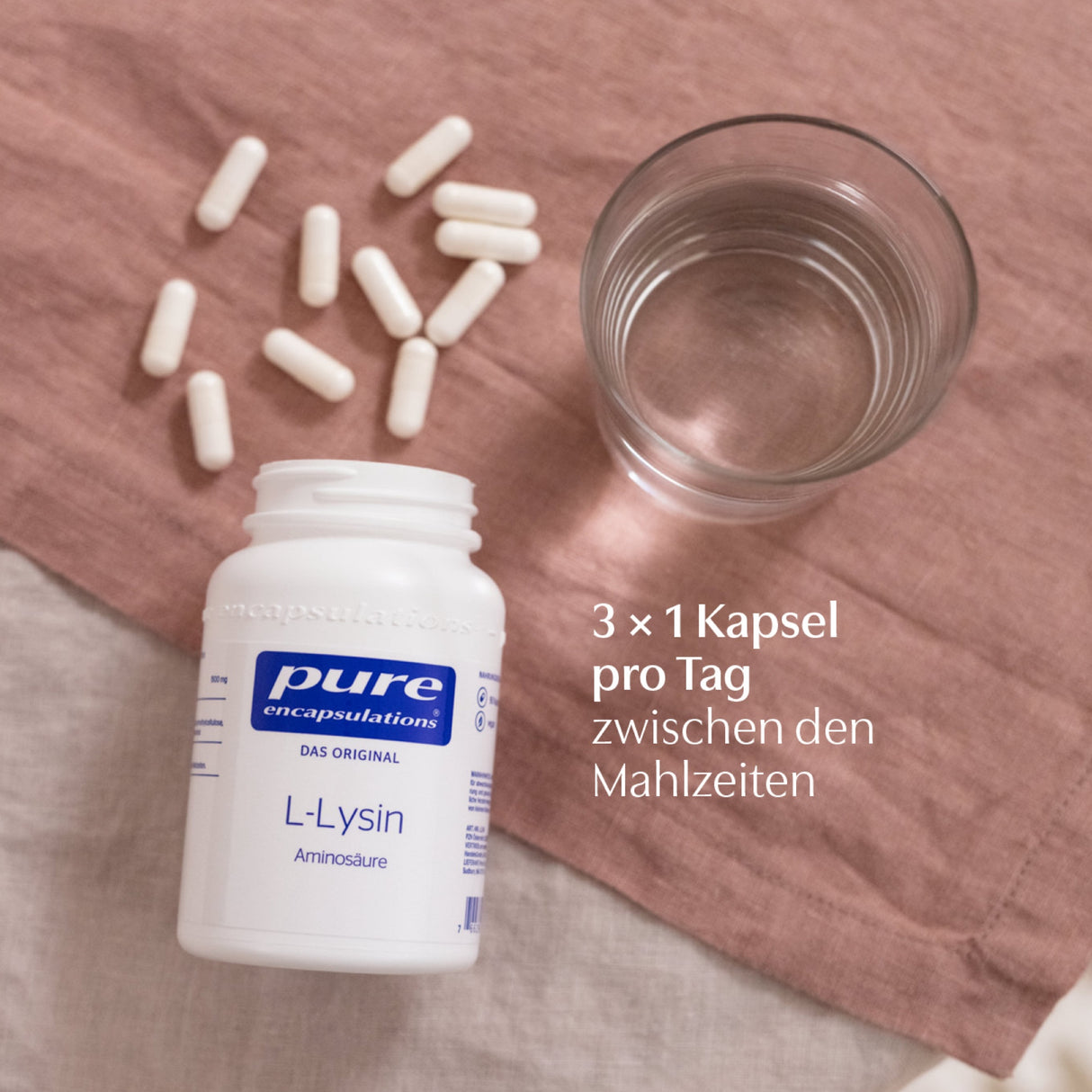 Ein geöffnetes Behältnis mit der Aufschrift 'pure encapsulations L-Lysin Aminosäure' liegt auf einem rosa Tuch. Daneben sind weiße Kapseln verstreut und ein Glas Wasser steht bereit. Ein Text empfiehlt die Einnahme von 3x1 Kapsel pro Tag zwischen den Mahlzeiten.