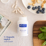 Eine offene weiße Dose mit der Aufschrift "pure encapsulations All-in-one Basisversorgung" liegt auf einem Tisch. Daneben liegen Kapseln, Blaubeeren und Nüsse auf einem Teller. Ein Holzbrett mit Spinatblättern ist ebenfalls zu sehen. Text auf dem Bild: "1-3 Kapseln täglich zum Essen".