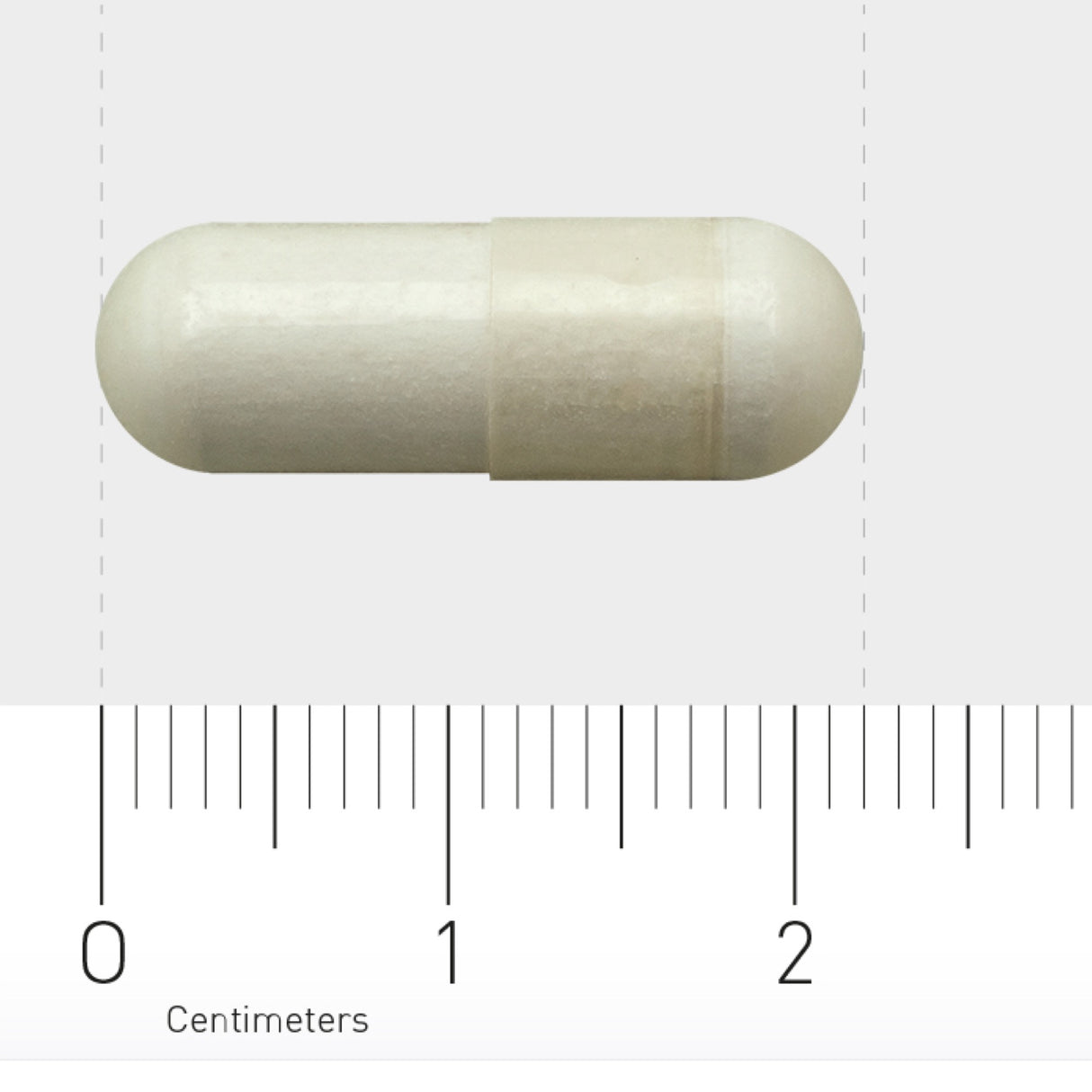 Das Bild zeigt eine weiße Kapsel auf einem Lineal mit Zentimeter-Maßstab. Die Kapsel ist zylindrisch und liegt horizontal. Der Maßstab zeigt, dass die Kapsel etwa 2 Zentimeter lang ist. Der Hintergrund ist einfarbig hellgrau.