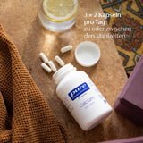 Ein geöffnetes weißes Fläschchen mit der Aufschrift 'Pure Encapsulations Calcium' liegt auf einem Tisch. Daneben liegen mehrere weiße Kapseln und ein Glas Wasser mit Zitronenscheibe. Ein braunes Handtuch ist ebenfalls sichtbar.