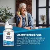 Eine ältere Frau mit langen weißen Haaren sitzt lächelnd an einem Tisch und hält einen Apfel in der Hand. Neben ihr steht eine Pflanze in einem weißen Topf. Im Vordergrund ist eine große Verpackung von Vitamin C 1000 Plus zu sehen, auf der die Produktbeschreibung steht. Der Hintergrund zeigt ein modernes Interieur mit einem Fenster und einem Ziegelwand-Design.