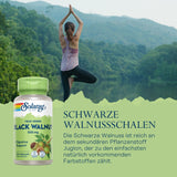 Eine Frau steht in Yoga-Pose vor einem See mit bewaldeten Hügeln im Hintergrund. Links im Bild ist eine Flasche Solaray True Herbs Black Walnut 500 mg zu sehen. Daneben steht der Text: 'SCHWARZE WALNUSSSCHALEN Die Schwarze Walnuss ist reich an dem sekundären Pflanzenstoff Juglon, der zu den einfachsten natürlich vorkommenden Farbstoffen zählt.'