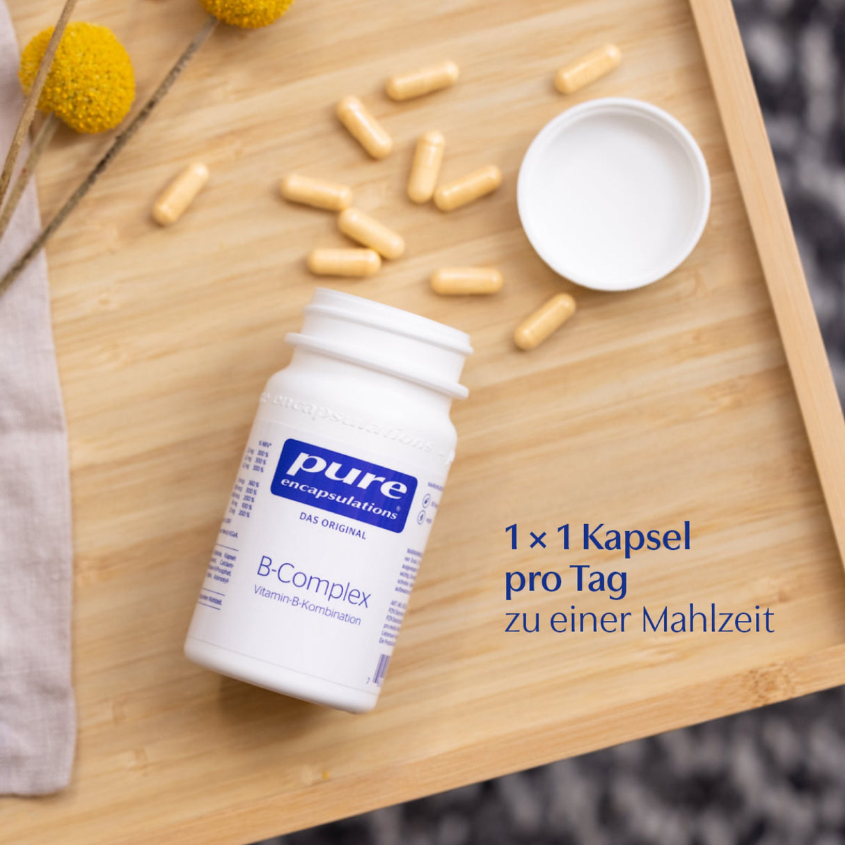 Das Bild zeigt eine weiße Flasche mit der Aufschrift 'pure encapsulations B-Complex Vitamin-B-Kombination' auf einem hölzernen Tablett. Die Flasche liegt auf der Seite, der Deckel ist abgenommen. Um die Flasche herum liegen mehrere gelbe Kapseln. Im Hintergrund sind gelbe Blumen zu sehen. Auf dem Bild steht in blauer Schrift: '1 x 1 Kapsel pro Tag zu einer Mahlzeit'.