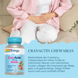 Das Bild zeigt eine Frau, die auf einem Sofa sitzt und ihre Hände schützend auf ihren Bauch legt. Sie trägt ein rosa Hemd und weiße Hosen. Im Vordergrund ist eine Flasche mit CranActin Chewables von Solaray zu sehen. Auf der Flasche steht, dass die Tabletten die Gesundheit der Harnwege unterstützen. Der Hintergrund ist hell und neutral gehalten. Der Text beschreibt die Vorteile von Cranberry-Bestandteilen für die Blasengesundheit.
