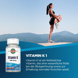 Das Bild zeigt eine Frau in Sportkleidung, die auf einem Felsen in den Bergen steht und eine Dehnübung macht. Im Vordergrund ist eine Flasche Vitamin K1 von KAL zu sehen. Der Hintergrund zeigt einen klaren blauen Himmel und Berglandschaft.