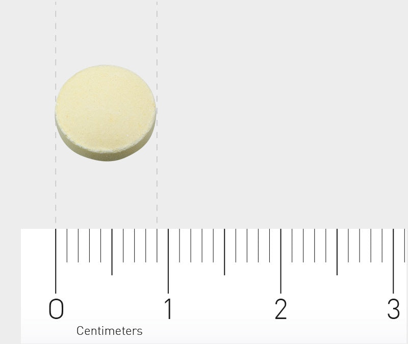 Das Bild zeigt eine einzelne, runde, cremefarbene Tablette auf einem grauen Hintergrund. Unter der Tablette befindet sich ein Lineal mit Zentimetereinteilung, das die Größe der Tablette veranschaulicht. Die Tablette ist etwa 1,5 cm im Durchmesser.