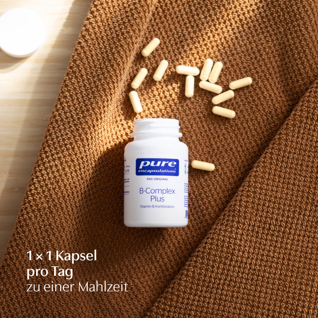Eine weiße Flasche mit der Aufschrift 'pure encapsulations B-Complex Plus' liegt auf einem braunen Stoff. Daneben sind mehrere gelbe Kapseln verstreut. In der linken unteren Ecke steht der Text '1 × 1 Kapsel pro Tag zu einer Mahlzeit'.