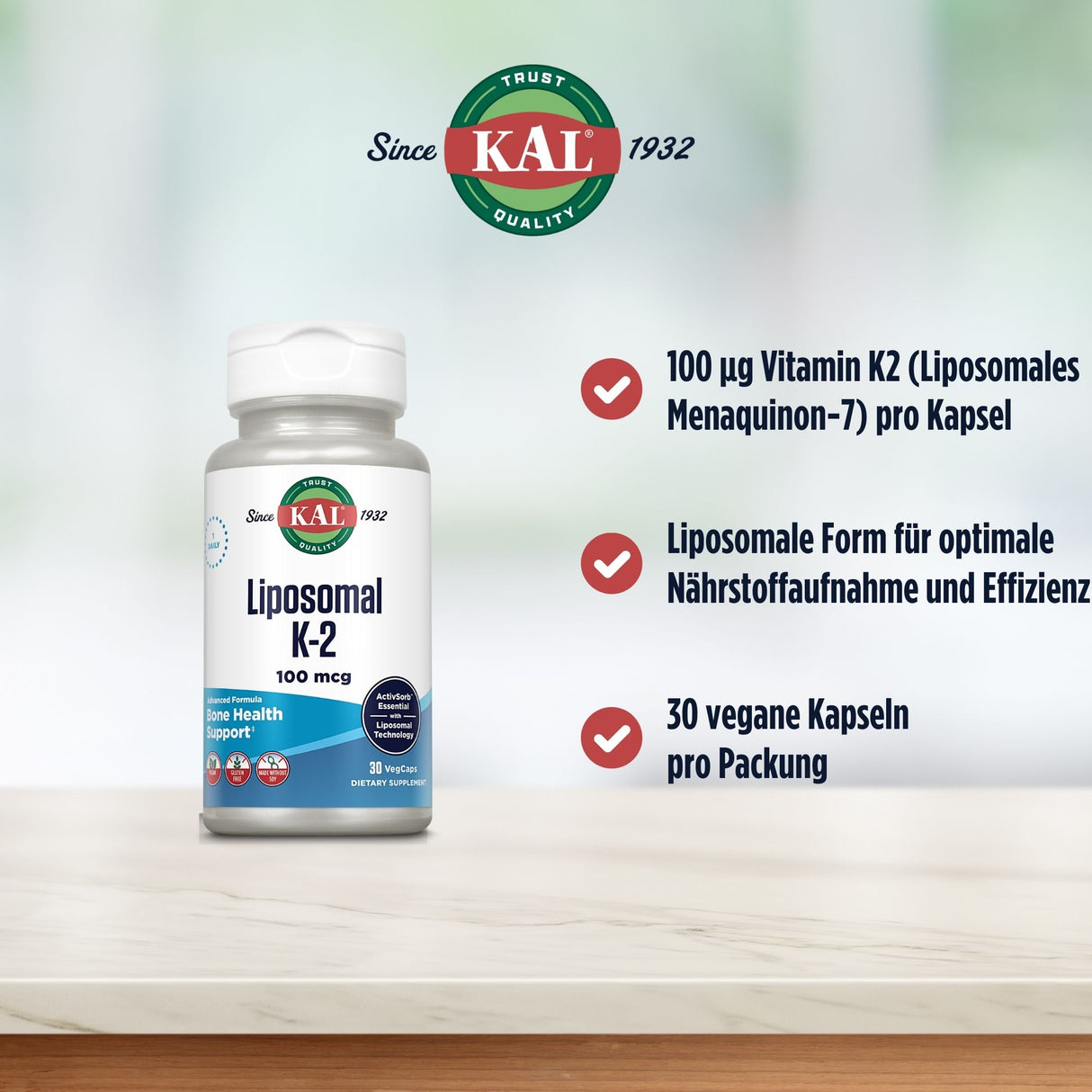 Das Bild zeigt eine Flasche KAL Liposomal K-2 Supplement auf einem Tisch. Die Flasche ist weiß mit einem Etikett, das den Produktnamen und die Dosierung von 100 mcg anzeigt. Daneben befinden sich drei Aufzählungspunkte: 100 µg Vitamin K2 (Liposomales Menaquinon-7) pro Kapsel, Liposomale Form für optimale Nährstoffaufnahme und Effizienz, 30 vegane Kapseln pro Packung. Im Hintergrund ist ein verschwommener, heller Hintergrund zu sehen. Das KAL-Logo ist oben links abgebildet mit dem Text "Since 1932".