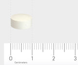 Das Bild zeigt eine weiße, runde Tablette, die auf einem Lineal mit Zentimeterangaben liegt. Die Tablette ist etwa 1 Zentimeter im Durchmesser. Der Hintergrund ist neutral und hellgrau.