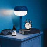 Ein blauer Nachttisch mit einer modernen blauen Tischlampe, einem klassischen Wecker, einer Flasche mit der Aufschrift 'RESTFUL YOU', einem Buch und einer Schlafmaske. Der Hintergrund ist ebenfalls blau.