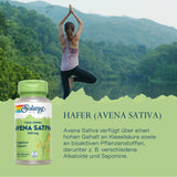 Eine Frau steht in der Yoga-Pose Vrksasana (Baumhaltung) vor einem See mit bewaldeten Hügeln im Hintergrund. Sie trägt ein lila Tanktop und schwarze Leggings. Im Vordergrund ist eine Verpackung von Solaray True Herbs Avena Sativa 350 mg abgebildet. Daneben steht der Text: 'Hafer (Avena Sativa)'. Darunter wird beschrieben, dass Avena Sativa Kieselsäure und bioaktive Pflanzenstoffe enthält, darunter Alkaloide und Saponine.