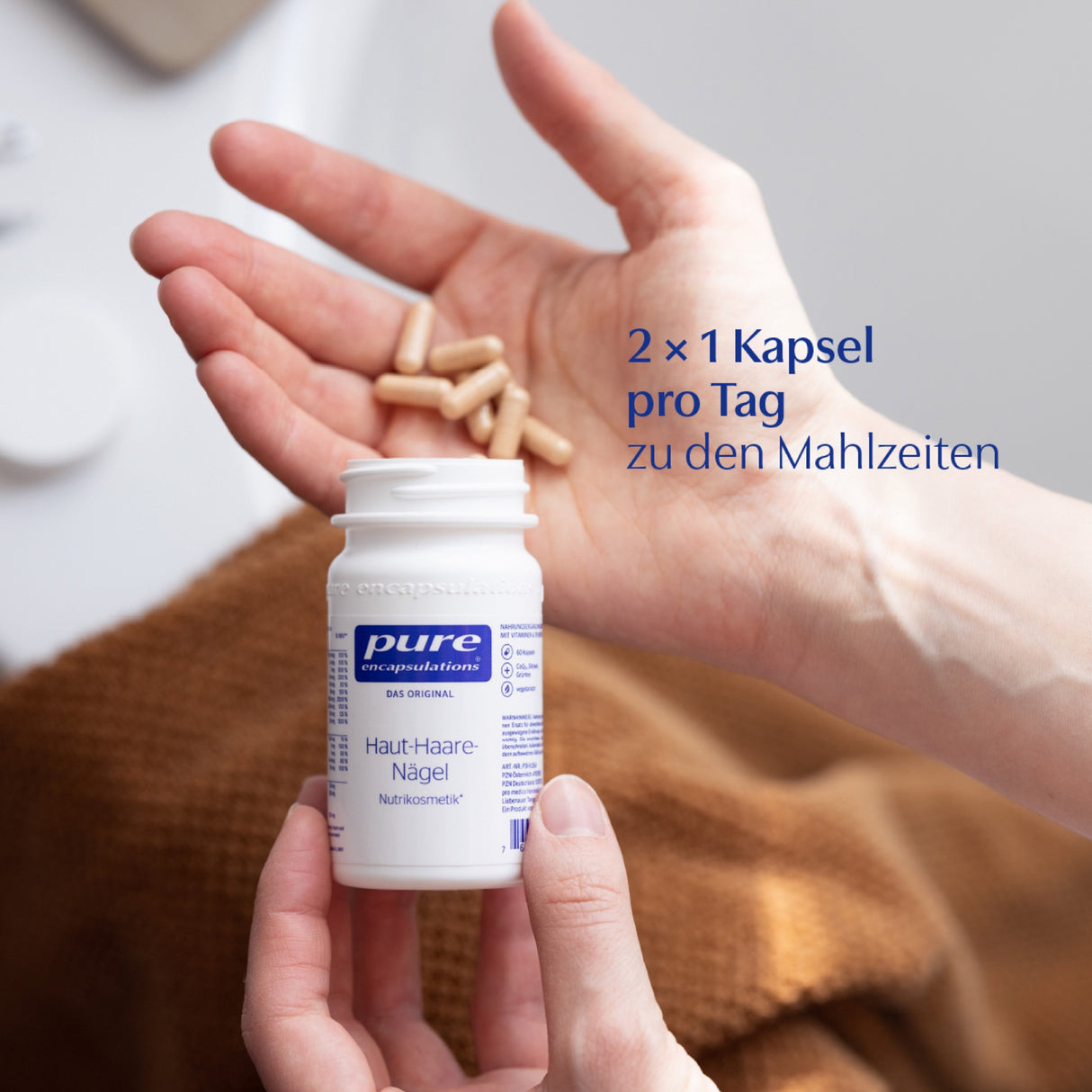 Eine Hand hält eine geöffnete Flasche "pure encapsulations" Nahrungsergänzungsmittel mit der Aufschrift "Haut-Haare-Nägel". In der anderen Hand sind mehrere Kapseln zu sehen. Im Hintergrund ist ein unscharfer, heller Untergrund. Auf dem Bild steht der Text "2 x 1 Kapsel pro Tag zu den Mahlzeiten".