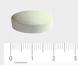 Eine weiße Tablette liegt auf einem Lineal, das in Zentimetern skaliert ist. Die Tablette hat einen Durchmesser von etwa 1,5 cm. Der Hintergrund ist neutral grau.