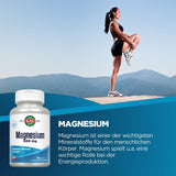 Eine Frau in Sportkleidung steht auf einem Felsen in einer Berglandschaft und dehnt ihr Bein. Im Vordergrund ist eine Flasche Magnesiumtabletten mit der Aufschrift 'Magnesium 500 mg' zu sehen. Der Hintergrund zeigt blauen Himmel und Berge.