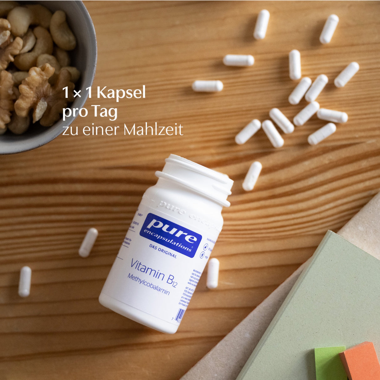 Das Bild zeigt eine weiße Flasche mit der Aufschrift 'pure encapsulations Vitamin B12 Methylcobalamin' auf einem Holztisch. Daneben liegen weiße Kapseln verstreut. Links oben ist eine Schale mit Nüssen zu sehen. Auf dem Bild steht der Text '1 x 1 Kapsel pro Tag zu einer Mahlzeit'.