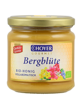 Ein Glas Hoyer Gourmet Bio-Honig 'Bergblüte' mit goldfarbenem Deckel und buntem Etikett mit Blumenmotiv.