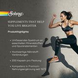 Das Bild zeigt eine sportliche Frau in Bewegung, die Sportbekleidung trägt. Links im Bild ist das Logo von Solaray zu sehen, darunter der Slogan 'SUPPLEMENTS THAT HELP YOU LIVE BRIGHTER'. Darunter stehen Produkthighlights wie 'Umfassendes Spektrum an essentiellen Mineralstoffen und Spurenelementen', 'Hochwertige Nährstoff-Verbindungen', '200 Kapseln pro Packung' und 'Kompetenz in Premium-Nahrungsergänzung seit 1973'. Der Hintergrund ist eine graue Betonwand.