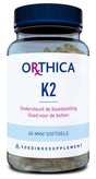 Ein Glasgefäß mit einem blauen Deckel enthält 60 Mini-Softgels eines Nahrungsergänzungsmittels namens Orthica K2. Der Text auf dem Etikett ist in Blau und Pink gehalten. Es wird beworben, dass das Produkt die Blutgerinnung unterstützt und gut für die Knochen ist.