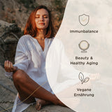 Eine Frau sitzt im Schneidersitz auf einem Felsen und meditiert. Sie trägt ein weißes Kleid. Neben ihr sind grafische Symbole und Texte zu den Themen Immunbalance, Beauty & Healthy Aging und Vegane Ernährung zu sehen.